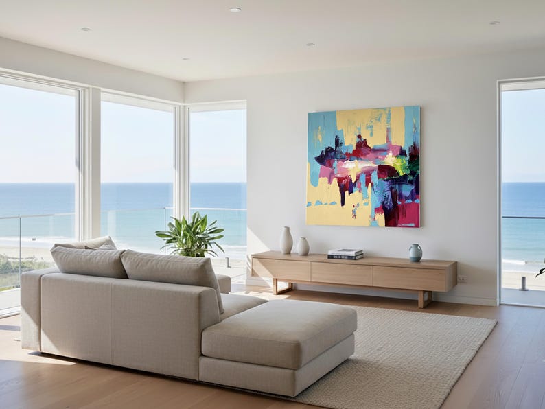 May include: A modern living room with a beige sectional sofa, a light wood media console, and a large abstract painting with blue, yellow, and red hues. The room has large windows overlooking the ocean.