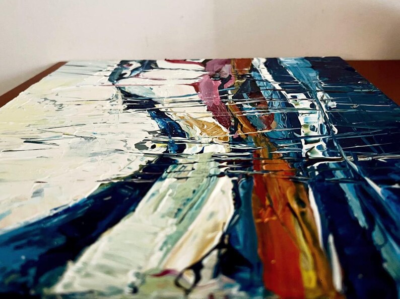 May include: Abstract painting with thick brushstrokes in shades of white, blue, orange, and pink. The artwork has a textured surface, creating a dynamic and expressive visual effect. The painting is on a square canvas.