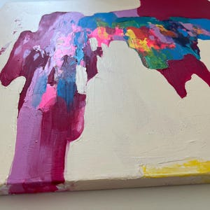 May include: Abstract canvas painting with a cream backdrop. The artwork features bold strokes of magenta, pink, blue, yellow, and green paint, creating a dynamic composition. The textured surface adds to the modern aesthetic.