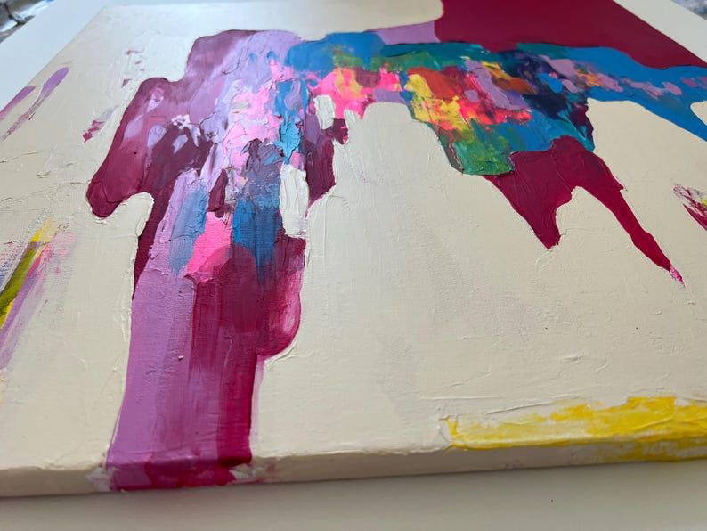May include: Abstract canvas painting with a cream backdrop. The artwork features bold strokes of magenta, pink, blue, yellow, and green paint, creating a dynamic composition. The textured surface adds to the modern aesthetic.
