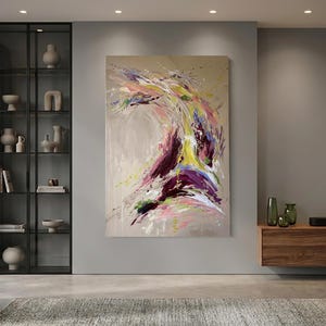 May include: Large abstract painting with a beige background and dynamic brushstrokes in shades of purple, yellow, pink, and green. The artwork is mounted on a gray wall in a modern living room setting.