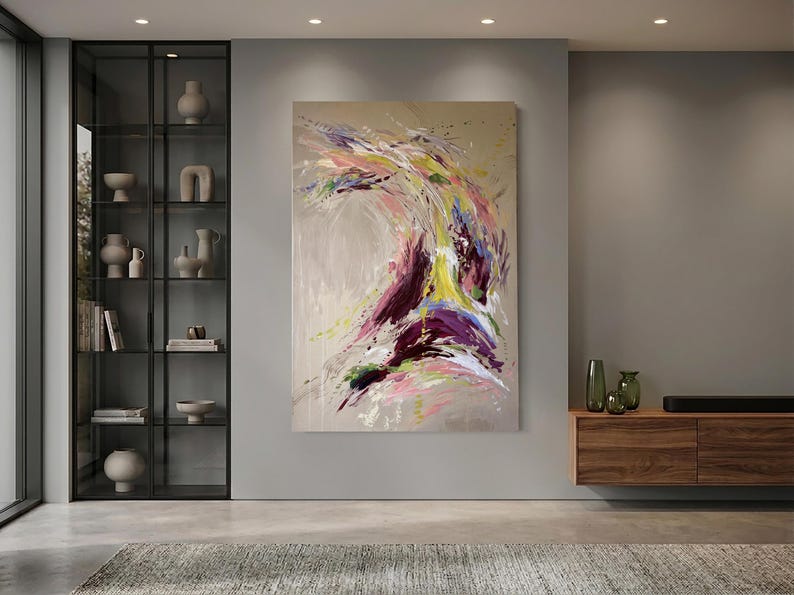 May include: Large abstract painting with a beige background and dynamic brushstrokes in shades of purple, yellow, pink, and green. The artwork is mounted on a gray wall in a modern living room setting.