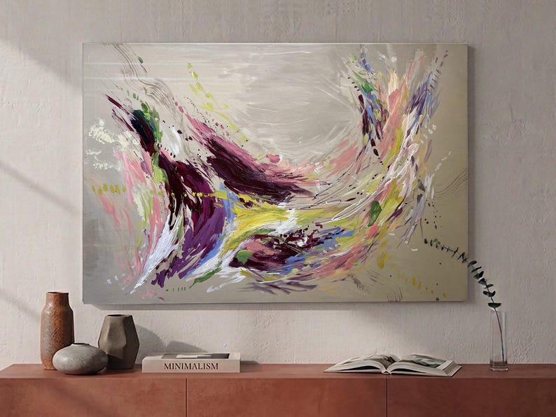 May include: Abstract painting with a neutral background and dynamic brushstrokes in shades of purple, pink, yellow, and green. The artwork features a burst of color and movement, creating a vibrant and energetic composition. The painting is displayed above a wooden cabinet with decorative vases and a book.