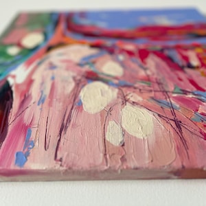 May include: Close-up of a square canvas with an abstract painting. The artwork features a textured surface with a palette of pink, red, blue, green, and white. Brushstrokes and layered paint create a dynamic composition.
