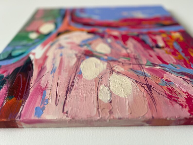May include: Close-up of a square canvas with an abstract painting. The artwork features a textured surface with a palette of pink, red, blue, green, and white. Brushstrokes and layered paint create a dynamic composition.