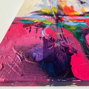 May include: Close-up of an abstract painting with thick brushstrokes. The artwork features a vibrant palette of pink, purple, orange, blue, and green hues. The textured surface of the canvas is visible, showcasing the artist's technique and use of color.