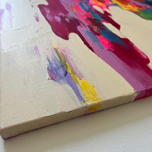 May include: Abstract canvas painting with textured brushstrokes. The artwork has a cream background with bold splashes of magenta, pink, yellow, blue, and green paint. The paint is layered, creating a dynamic visual effect. The canvas is approximately 2.5 cm thick.