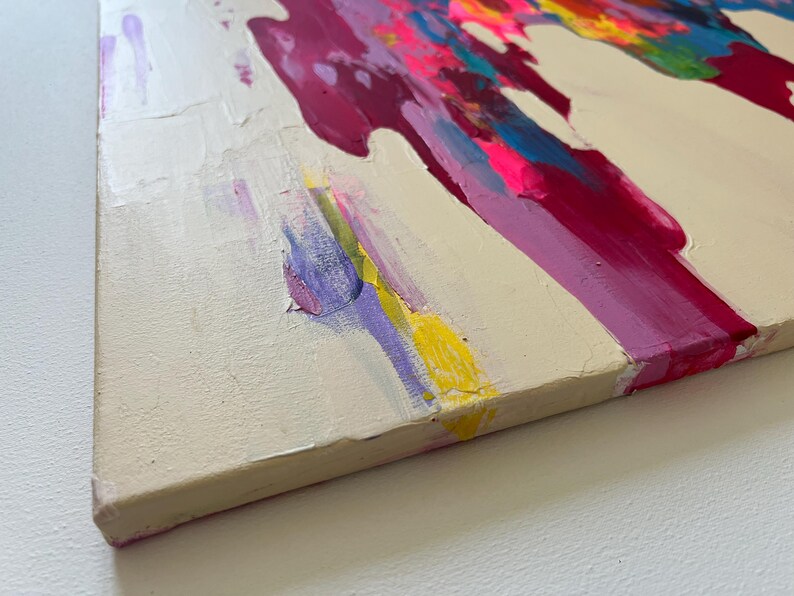 May include: Abstract canvas painting with textured brushstrokes. The artwork has a cream background with bold splashes of magenta, pink, yellow, blue, and green paint. The paint is layered, creating a dynamic visual effect. The canvas is approximately 2.5 cm thick.