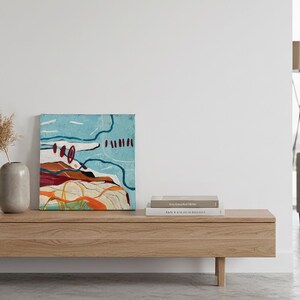 May include: An abstract painting with a blue sky and earth-toned landscape sits on a wooden console. A vase with dried pampas grass and a stack of books complete the minimalist decor. The artwork features red, orange, and white accents.