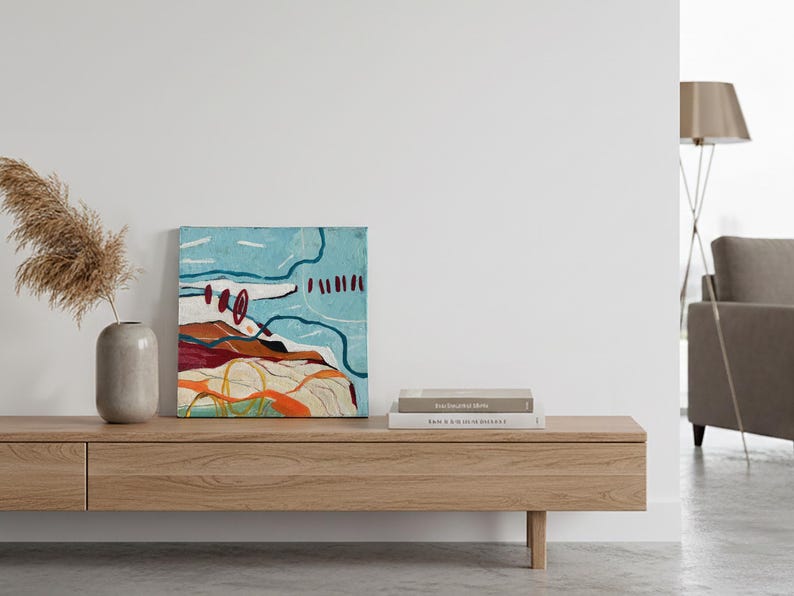 May include: An abstract painting with a blue sky and earth-toned landscape sits on a wooden console. A vase with dried pampas grass and a stack of books complete the minimalist decor. The artwork features red, orange, and white accents.