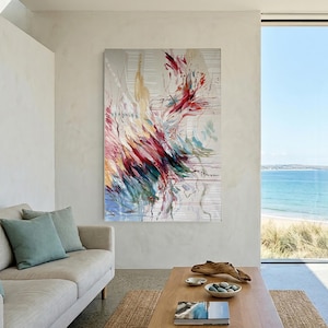 May include: Abstract art piece with a vibrant display of colors, including red, yellow, and blue, against a neutral background. The artwork is displayed in a modern living room setting, complemented by a neutral-toned sofa and a view of the ocean.