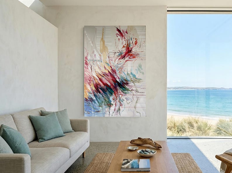 May include: Abstract art piece with a vibrant display of colors, including red, yellow, and blue, against a neutral background. The artwork is displayed in a modern living room setting, complemented by a neutral-toned sofa and a view of the ocean.