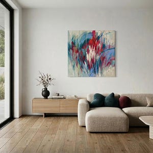 May include: An abstract painting with red, blue, and white brushstrokes hangs above a beige sectional sofa. The room features a wooden floor, a low wooden cabinet, and a black vase with branches. Large windows offer a view of the outdoors.