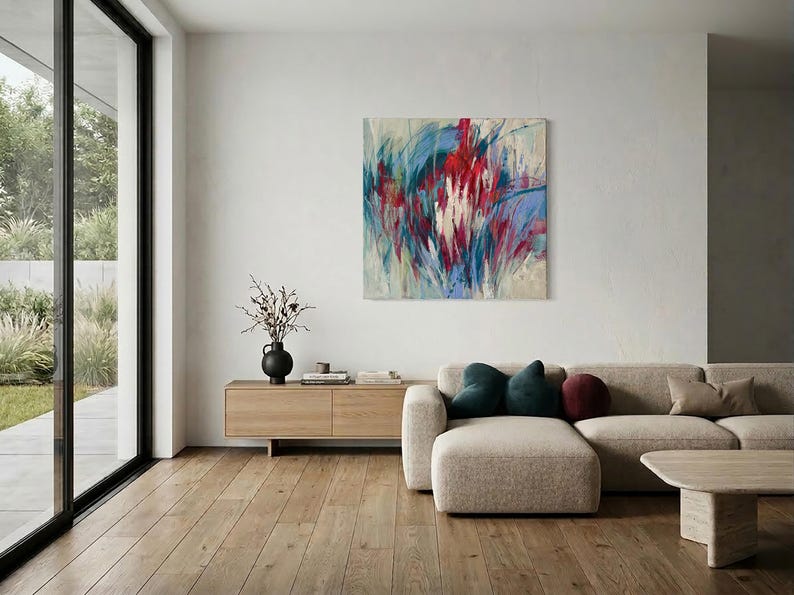 May include: An abstract painting with red, blue, and white brushstrokes hangs above a beige sectional sofa. The room features a wooden floor, a low wooden cabinet, and a black vase with branches. Large windows offer a view of the outdoors.