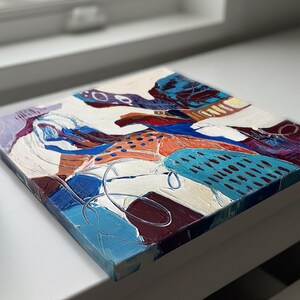 以下が含まれることがあります： 正方形の抽象絵画で、テクスチャ加工された表面です。この作品は、白、青、オレンジ、紫など、さまざまな色が混ざっています。構成はさまざまな形とパターンで構成されており、筆致が見え、塗料が厚く塗られています。