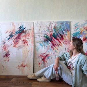 May include: Three abstract paintings with vibrant brushstrokes in shades of red, blue, and green. The artwork is displayed against a white background. A person is sitting on the floor next to the paintings.