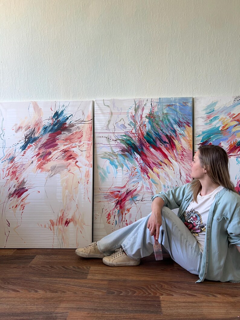 May include: Three abstract paintings with vibrant brushstrokes in shades of red, blue, and green. The artwork is displayed against a white background. A person is sitting on the floor next to the paintings.