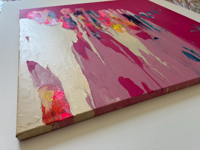 May include: Abstract painting on canvas with a vibrant color palette. The artwork features a combination of pink, white, yellow, and blue hues, with paint drips and textured brushstrokes. The canvas is stretched and mounted on a wooden frame.