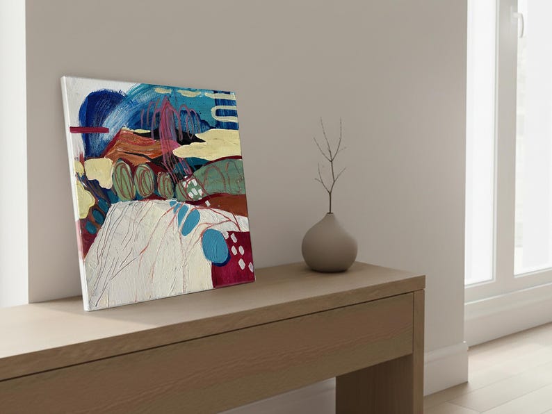 May include: Abstract art canvas with a mix of colors including blue, red, yellow, and green. The artwork features brushstrokes and shapes, creating a modern aesthetic. A small, gray vase with a twig is next to the canvas.