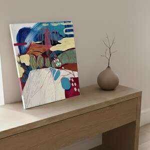 May include: An abstract painting on canvas featuring a landscape with blue, yellow, and red brushstrokes. The artwork is displayed on a light-colored wooden surface, next to a small, round, gray vase with a twig.