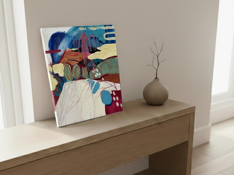 May include: An abstract painting on canvas featuring a landscape with blue, yellow, and red brushstrokes. The artwork is displayed on a light-colored wooden surface, next to a small, round, gray vase with a twig.