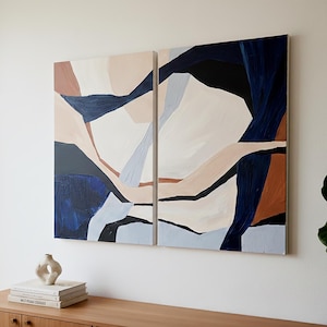 May include: Two abstract paintings with a modern design. The artwork features geometric shapes in shades of navy blue, beige, brown, and white. The paintings are mounted on a white wall above a wooden cabinet.