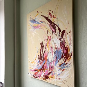 May include: Abstract painting on canvas with a beige background. The artwork features dynamic brushstrokes in shades of burgundy, pink, blue, yellow, and white. The painting is mounted on a wooden frame and displayed on a light green wall.