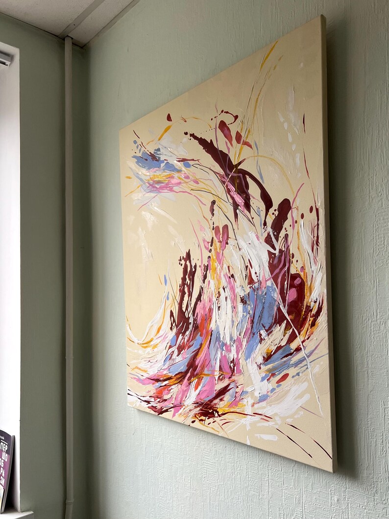 May include: Abstract painting on canvas with a beige background. The artwork features dynamic brushstrokes in shades of burgundy, pink, blue, yellow, and white. The painting is mounted on a wooden frame and displayed on a light green wall.