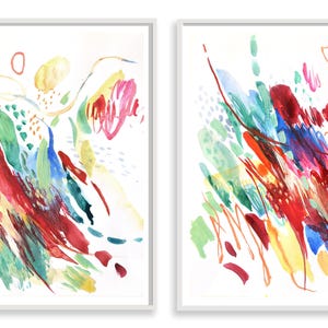 May include: Two framed vertical wall art prints featuring abstract watercolor paintings. The compositions display energetic brushstrokes and splatters in vibrant shades of red, blue, yellow, and green against a clean white background.