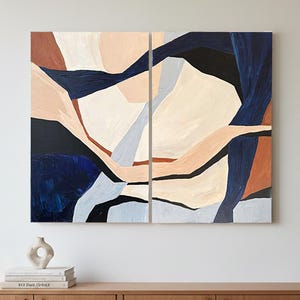 Op de afbeelding: Abstract kunstwerk in twee panelen. Het schilderij toont geometrische vormen in marineblauw, beige, lichtbruin en gebroken wit. Het kunstwerk is gemonteerd op een witte muur boven een houten kast.