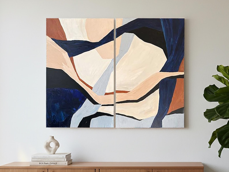 May include: Abstract art piece in two panels. The artwork features a composition of geometric shapes in shades of navy blue, beige, light brown, and off-white. The painting is mounted on a white wall above a wooden cabinet.
