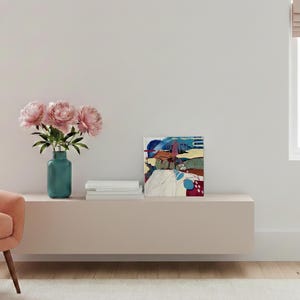 May include: A modern living room scene featuring a pink armchair, a teal vase with pink peonies, and an abstract painting. The painting has blue, red, and yellow hues. A stack of white books sits next to the painting.