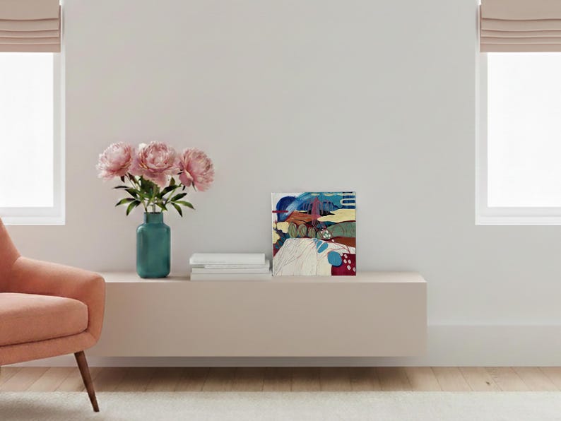 May include: A modern living room scene featuring a pink armchair, a teal vase with pink peonies, and an abstract painting. The painting has blue, red, and yellow hues. A stack of white books sits next to the painting.