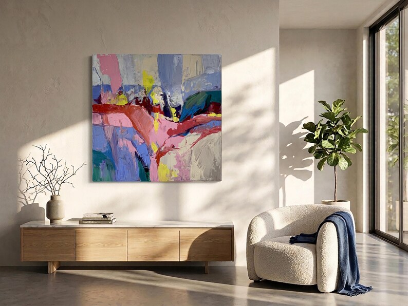 May include: Abstract oil painting with a vibrant colour palette. The artwork features bold brushstrokes in shades of pink, blue, yellow, and red. The painting is displayed on a wall above a wooden cabinet. A modern living room setting with a white chair and a plant.