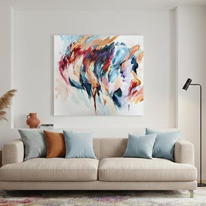 May include: An abstract painting in shades of blue, orange, and purple hangs above a beige sofa with throw pillows. A wooden side table holds a blue vase with dried plants. A black floor lamp stands to the right.