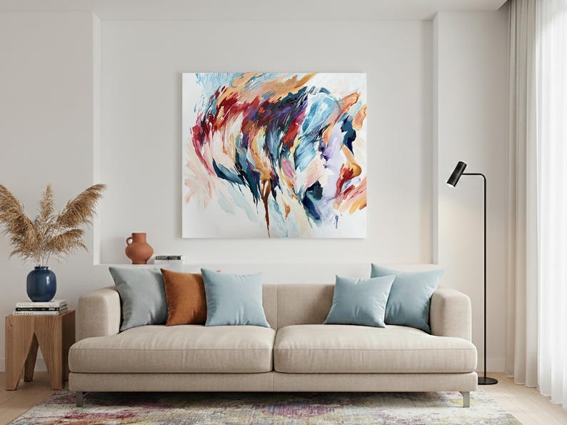May include: An abstract painting in shades of blue, orange, and purple hangs above a beige sofa with throw pillows. A wooden side table holds a blue vase with dried plants. A black floor lamp stands to the right.