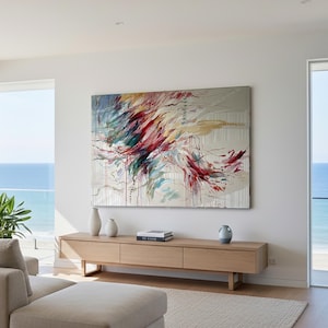 May include: Large abstract painting with red, teal, and yellow brushstrokes on a neutral background. The artwork hangs above a light wood console table with decorative vases and books. The room has large windows overlooking the ocean.