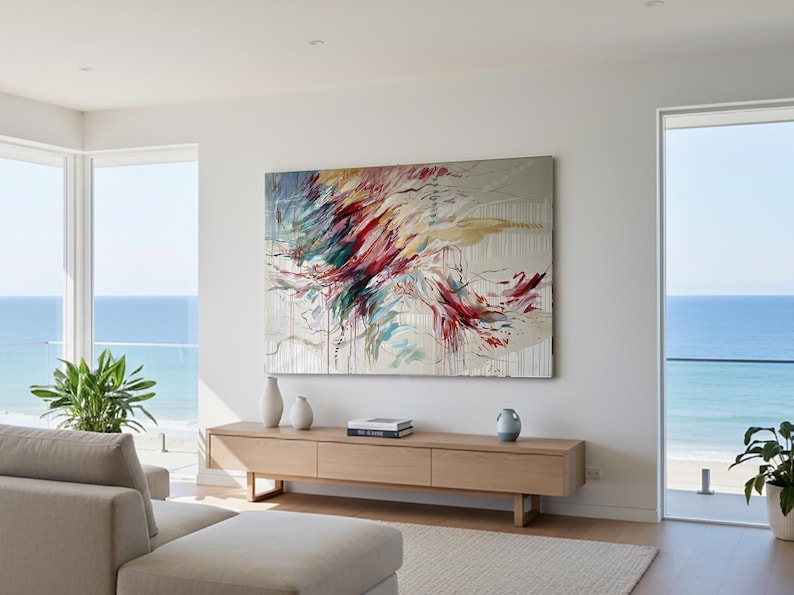 May include: Large abstract painting with red, teal, and yellow brushstrokes on a neutral background. The artwork hangs above a light wood console table with decorative vases and books. The room has large windows overlooking the ocean.