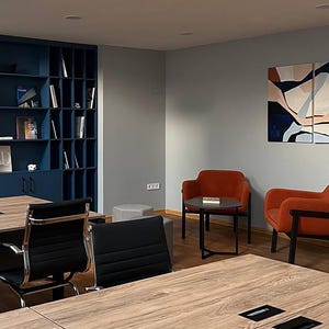 May include: An office interior featuring a modern design. The space includes a dark blue built-in bookshelf filled with books, two orange armchairs, and a small round table. Large wooden tables and black chairs are in the foreground.