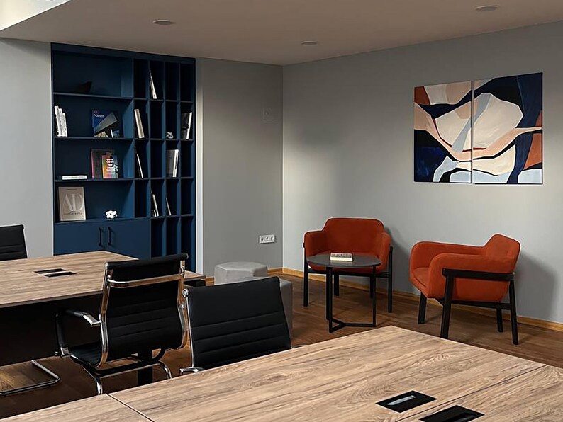 May include: An office interior featuring a modern design. The space includes a dark blue built-in bookshelf filled with books, two orange armchairs, and a small round table. Large wooden tables and black chairs are in the foreground.