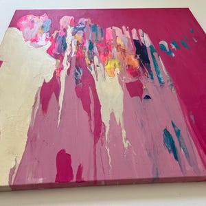 May include: Abstract painting on canvas with a vibrant color palette. The artwork features a bold fuchsia background with gold leaf accents and colorful drips of paint in shades of pink, blue, yellow, and white. The canvas is square and mounted on a wooden frame.