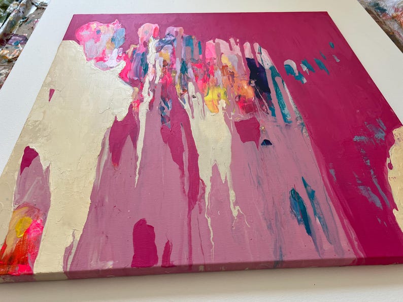 May include: Abstract painting on canvas with a vibrant color palette. The artwork features a bold fuchsia background with gold leaf accents and colorful drips of paint in shades of pink, blue, yellow, and white. The canvas is square and mounted on a wooden frame.
