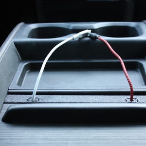 May include: A black car console with two cup holders and a small tray. A white USB cable and a red USB cable are plugged into the console. The console is made of black plastic and has a textured surface.