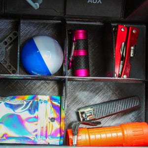 May include: A collection of items organized in a car console. Includes a beach ball, a multi-tool, a flashlight, a small binocular, a nail clipper, and a silver and black gear-like object. A holographic pouch is also visible.