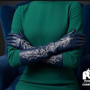 Puede incluir: Guantes largos de cuero azul marino con intrincado bordado floral blanco. Los guantes se extienden más allá de los codos y se usan con un vestido verde. Un llamativo accesorio de moda.