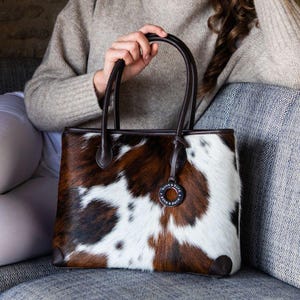 Może przedstawiać: Prostokątna torba typu tote z krowiej skóry w kolorze brązowym, białym i czarnym, z ciemnobrązowymi skórzanymi uchwytami i wykończeniami. Torba posiada mały okrągły charms z tekstem. Torba jest trzymana.