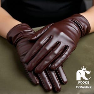 Puede incluir: Un par de guantes de cuero marrón oscuro. Los guantes son lisos y tienen un diseño clásico. La imagen muestra los guantes con las manos cruzadas.