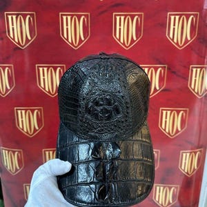 May include: Black baseball cap with a textured, crocodile-like pattern. The cap features a curved bill and a structured crown. The cap is held up against a red background with gold logos.