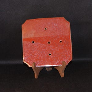 Peut inclure: Un porte-savon rectangulaire en céramique rouge avec cinq trous de drainage. Le plat a une texture mouchetée et est présenté sur un petit support en bois. Les bords du plat sont légèrement arrondis.