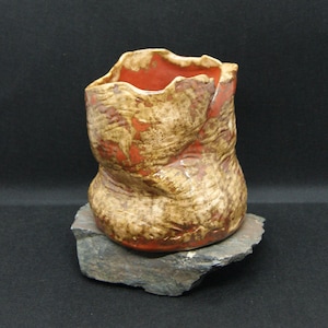 Peut inclure: Un vase en céramique fait à la main avec un design texturé et organique. Le vase présente des teintes marron, beige et rouge, avec une surface rugueuse et inégale. L'intérieur est rouge vif. Le vase est présenté sur une pierre grise.